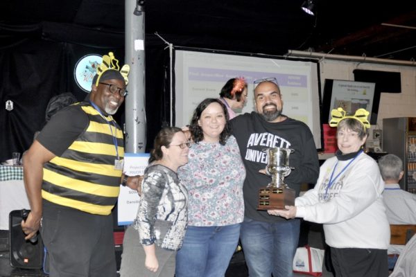 A group of four people, including one in a bee costume, smiling while holding a trophy cup at the Project Literacy JeopardBee event. A scoreboard is visible in the background as they celebrate their achievement.
