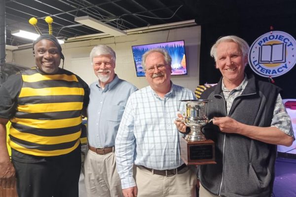Four men standing together, one in a bee costume, holding a silver trophy cup at the Project Literacy JeopardBee event. They are smiling, with the Project Literacy logo and event setting visible in the background.