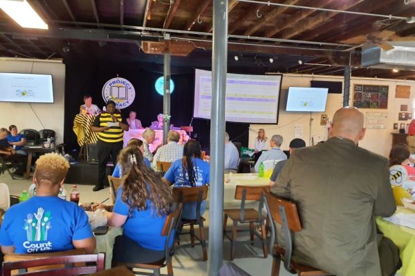 A broad view of the Project Literacy JeopardBee event, with attendees seated at tables while a person in a bee costume and others are on stage, managing the event and displaying the Jeopardy-style scoreboard on screens.