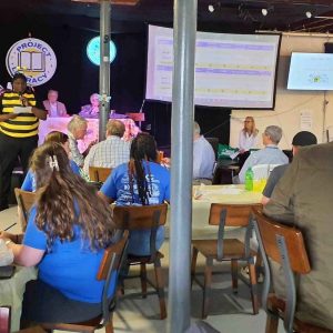 “View from the audience at a Project Literacy JeopardyBee event, showing attendees seated at tables while a host in a bee costume presents, with large screens displaying scores.”