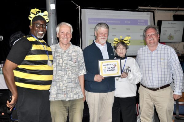 A group of five people standing together at the Project Literacy JeopardBee event, with one person dressed in a bee costume. Another person holds a framed certificate that reads “JeopardBee 2025 2nd Place Team Award.” They are smiling, with a projected scoreboard visible in the background.