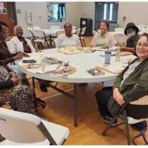 “A diverse group of adults sits around a round table in a gym setting, participating in a Project Literacy Book Buds discussion with books and snacks on the table.”