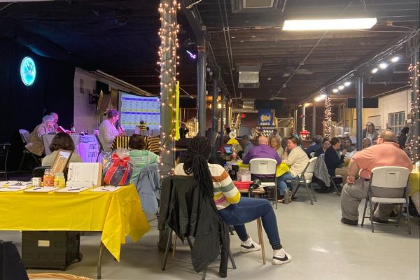A wide view of the Project Literacy JeopardBee event with attendees seated at tables under string lights, watching the stage where the game is projected. Participants are seen working on papers, and the room has a warm, community-focused atmosphere.