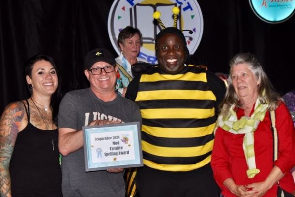 A group of four people at the Project Literacy JeopardBee event, one in a bee costume, smiling while one person holds a framed certificate that reads “JeopardBee 2024 Most Creative Spelling Award.” The Project Literacy logo is displayed in the background.