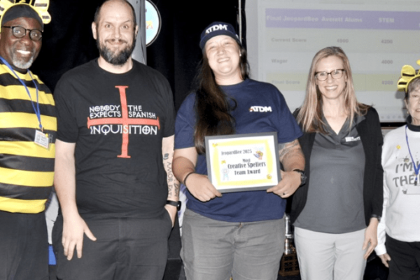 A group of five people stand together at a Project Literacy JeopardBee event. One is dressed in a bee costume, and another wears a "Nobody Expects the Spanish Inquisition" shirt. A woman holds a framed "Most Creative Spellers Team Award" plaque, with a screen and bee-themed décor in the background.
