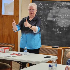 “An instructor with short white hair gives two thumbs up while teaching an American Sign Language class in front of a blackboard with written instructions and a welcome message.”