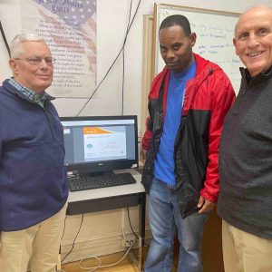 “Three men stand beside a computer displaying a certificate of achievement, smiling while participating in a Project Literacy computer literacy or digital learning session.”