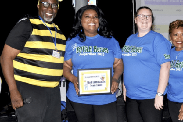 A group of five people at a Project Literacy JeopardBee event, with one in a bee costume and three women in matching blue "SOVAH Heroes" shirts. One woman holds a framed "Most Enthusiastic Team Award" while they all smile, with a scoreboard projected behind them.