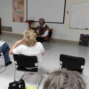 A group of adults sit and watch a woman at the front of a class room.