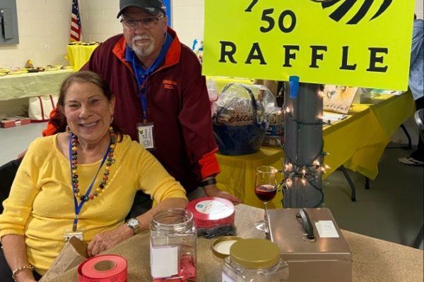 A woman seated and a man standing behind her at a table marked “50/50 Raffle” with a bright yellow sign and a cartoon bee. They are smiling, with jars for tickets and raffle items on the table during the Project Literacy JeopardBee event.