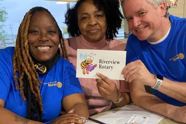 Three people seated at a table smiling, holding a sign that says “Riverview Rotary” with a cartoon bee reading a book. Two people are wearing blue Rotary shirts, and there are papers and snacks on the table in front of them.