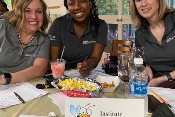 Three women seated at a table during an event, smiling while working on papers with snacks and drinks. A sign on the table reads “Institute for Advanced Learning & Research” with a bee reading a book.