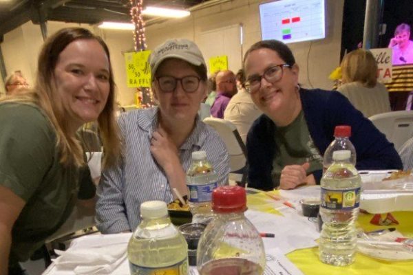 Three women seated at a table covered in a yellow tablecloth, smiling at the camera during an event. The table has food containers, water bottles, and a sign reading “Dog-Eared Page Sponsored by Infinity Global.”