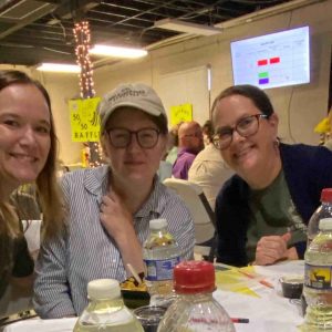 “Three smiling women seated together at a table during a Project Literacy JeopardyBee event, with bottles, raffle signs, and a presentation screen in the background.”