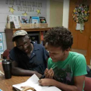 “An older man and a young man sit side by side, smiling while reading a book together during a Project Literacy tutoring session.”