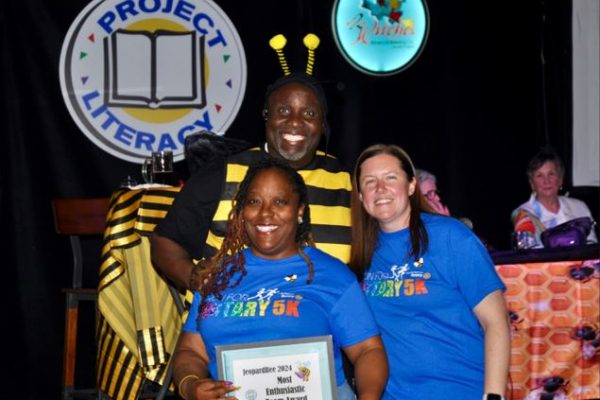 Three people smiling in front of a Project Literacy logo, with one person wearing a bee costume. They are holding a framed certificate for “Most Enthusiastic Team Award” at an event.