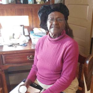 “An older woman wearing glasses, a pink sweater, and a black bonnet sits in front of a wooden desk, holding a small item while looking toward the camera.”
