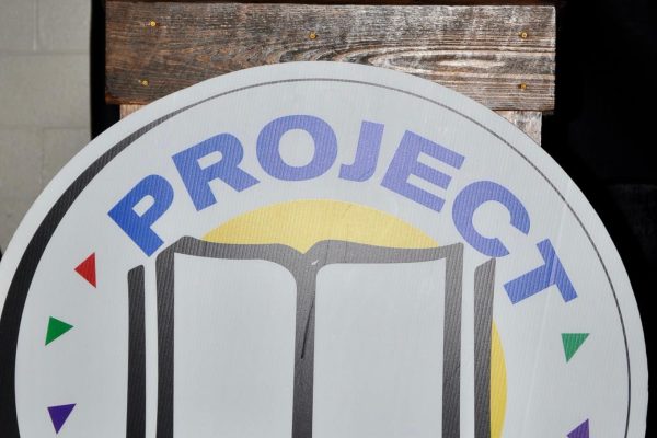 Woman smiles from behind a podium with a large Project Literacy logo sign during the JeopardBee event.